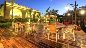 An outdoor dining area with wooden tables and chairs, a covered seating area, and a building with arched windows in the background. - The Westlake Hotel & Resort Yogyakarta (Sleman)