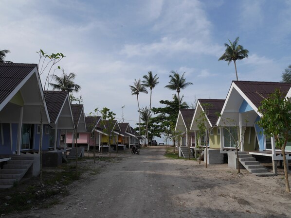 A row of colorful, single-story beach huts with a dirt road in between, surrounded by palm trees.