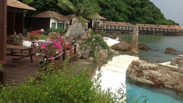 Un resort con bungalós sobre el agua, una terraza de madera con mobiliario exterior y vistas al océano y vegetación exuberante.