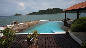 Una terraza de madera con piscina, rodeada por una costa rocosa y una isla distante.