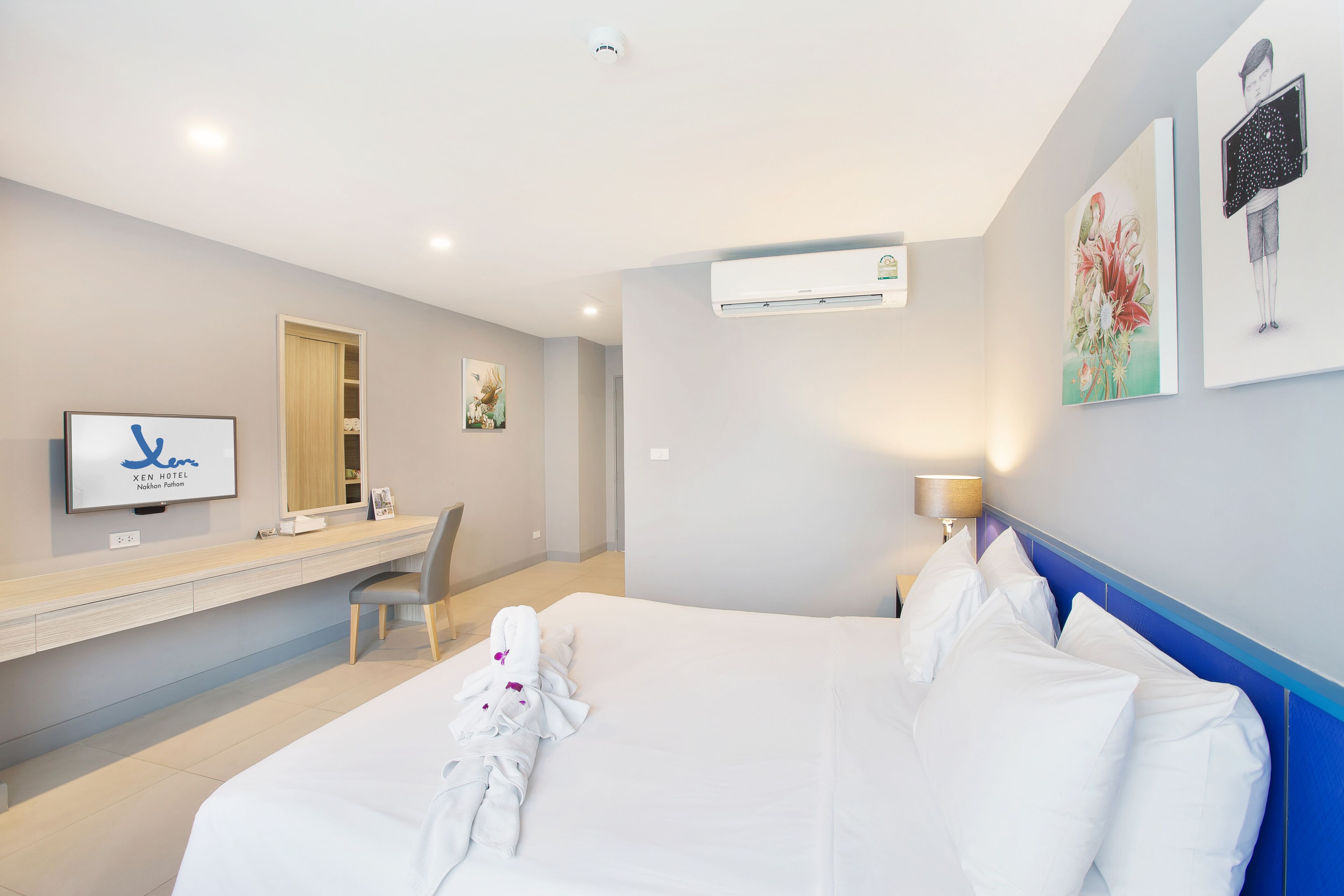 A hotel room with a bed, a desk, a chair, a TV, and artwork on the walls.
