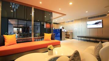A modern hotel lobby with a large seating area, a reception desk, and a TV screen.