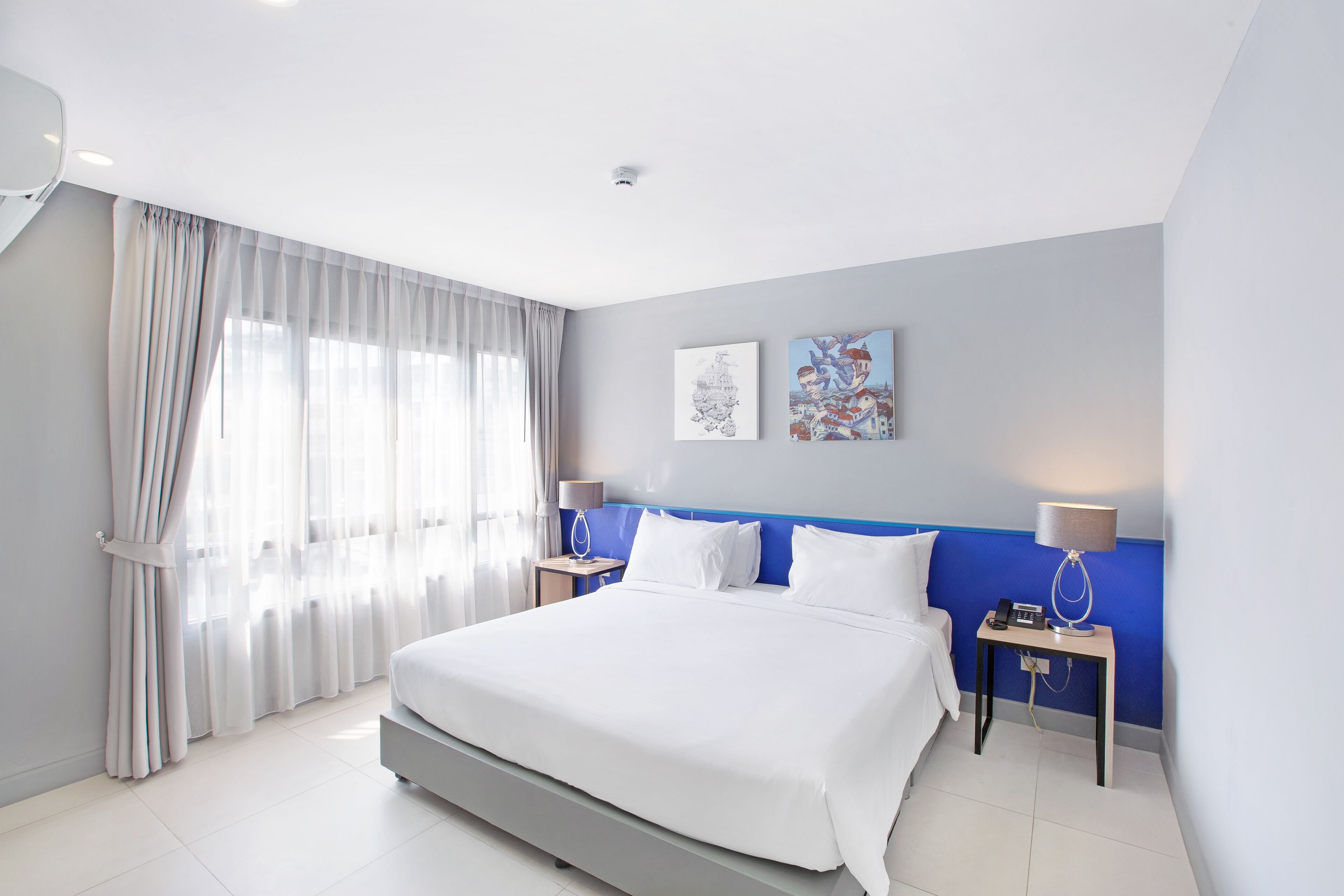 Une chambre d’hôtel moderne avec un grand lit, deux tables de chevet avec des lampes, une fenêtre avec des rideaux légers et deux œuvres d’art murales.