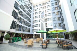 Un edificio moderno con varios pisos, zona de estar exterior con sillas y sombrillas, y un reloj digital grande.