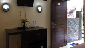 A room with a dark wooden desk, a flat-screen TV mounted on the wall, two wall-mounted lights, a door with a glass panel, and a small vase with a flower on the desk. - Jenny Bunker Homestay (Kuta)