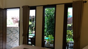 A hotel room with a bed, a small table, and a view of a garden through a window with curtains. - Jenny Bunker Homestay (Kuta)