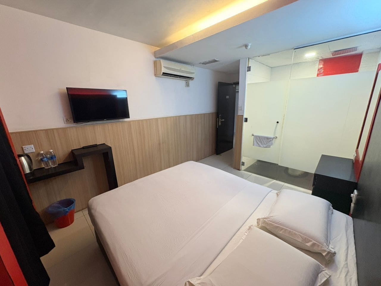 Ein Hotelzimmer mit Bett, Fernseher, Klimaanlage, einem kleinen Tisch mit Flaschen und einem Mülleimer.