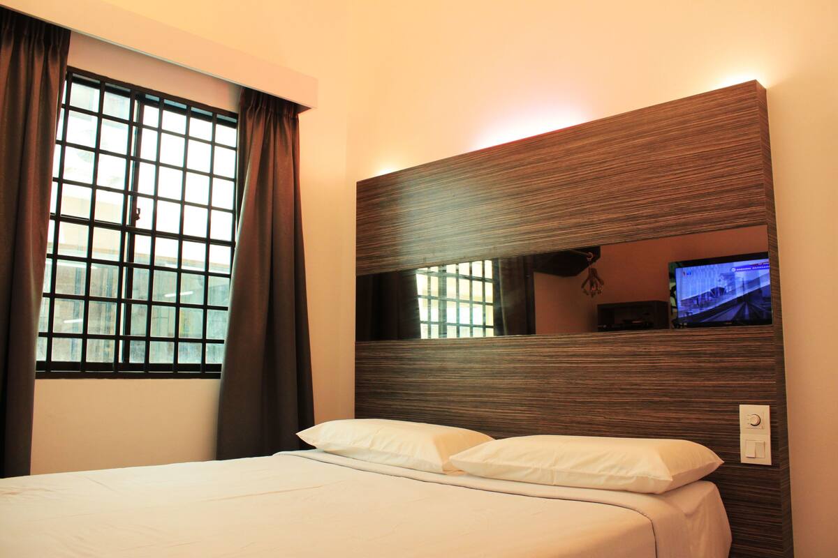 A hotel room with a bed, a window with curtains, a wall-mounted TV, and a wall switch.