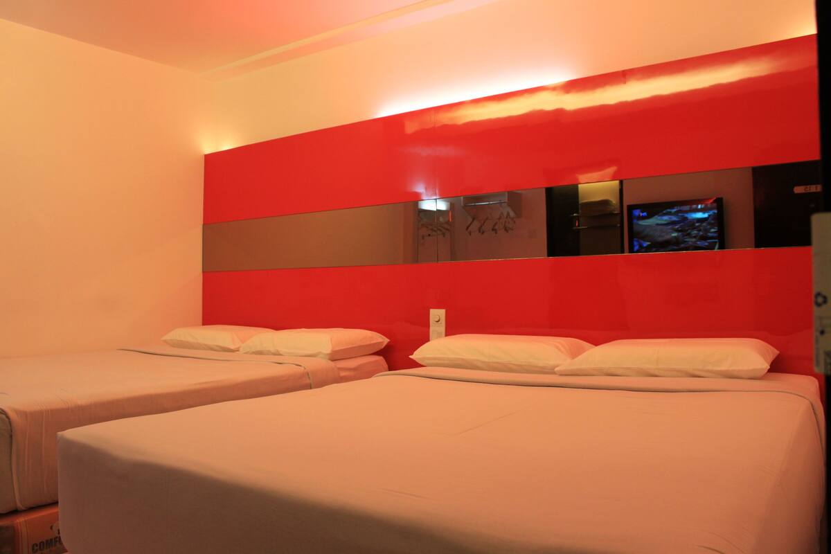 A hotel room with two beds, a red accent wall, and a television on the wall.