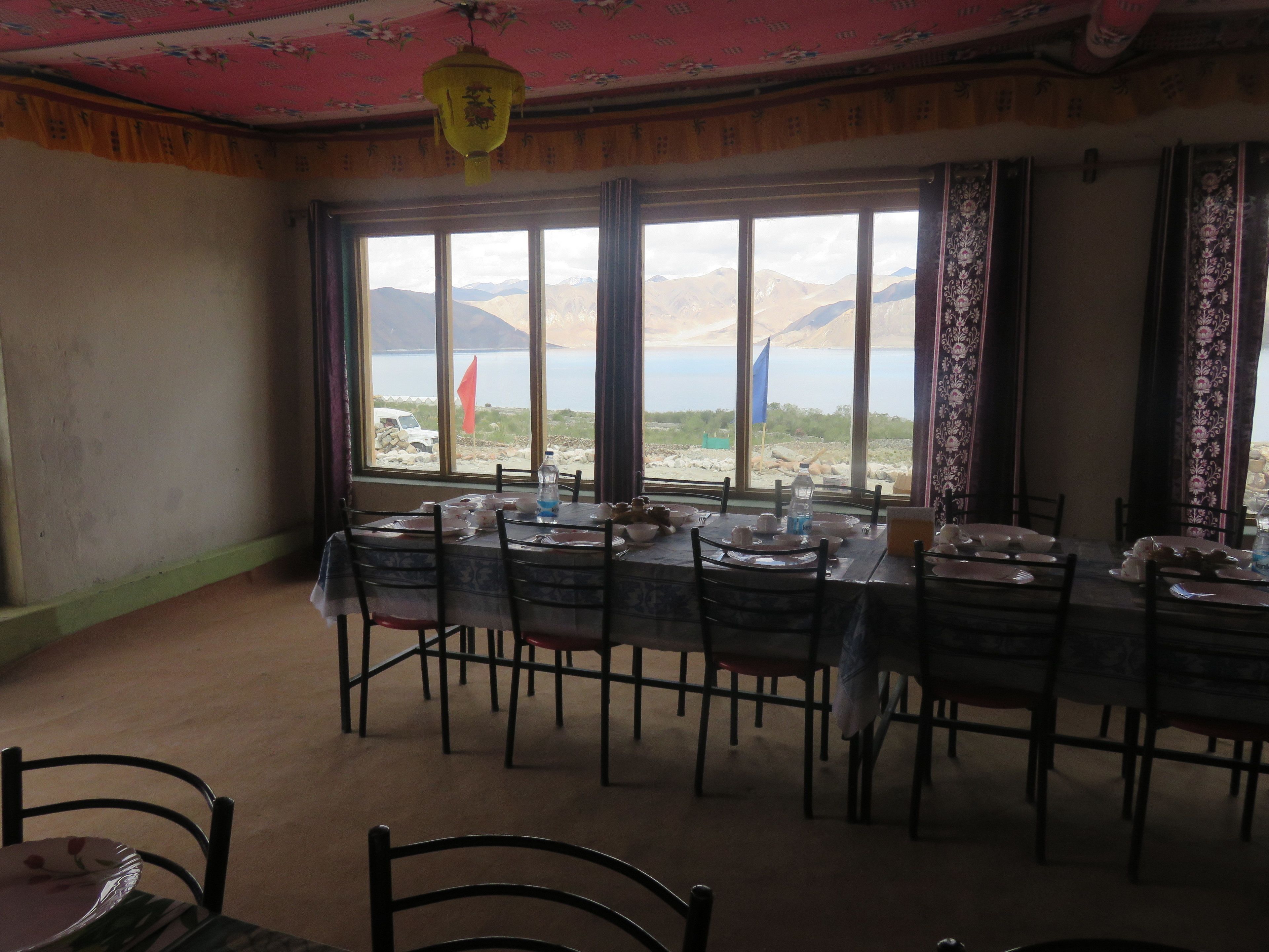 Un coin repas avec une table dressée pour six personnes, de grandes fenêtres offrant une vue sur les montagnes et un lac, et une lampe suspendue de style traditionnel.