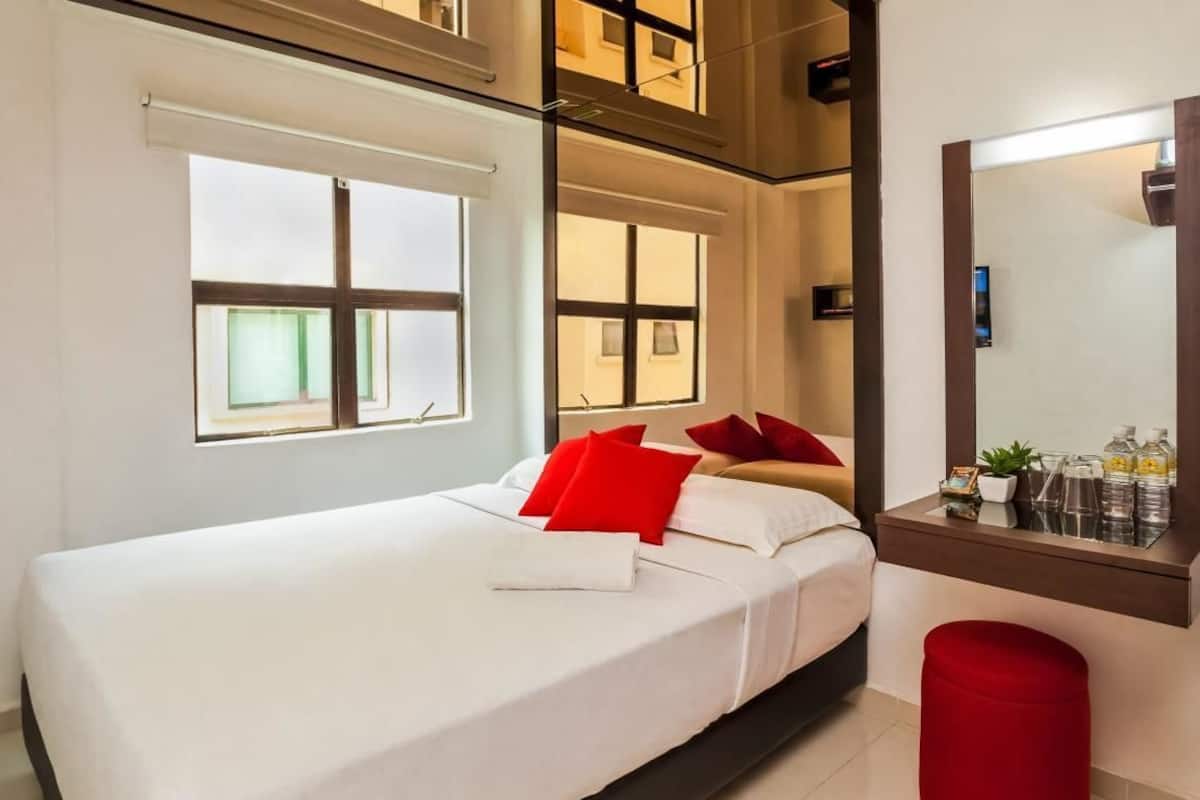 A modern hotel room with a bed, a red ottoman, a large mirror, and a window with blinds.