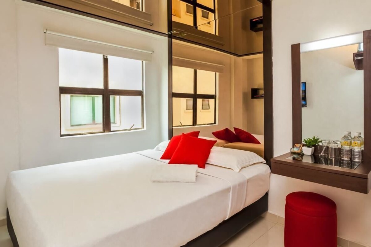 Una habitación de hotel moderna con una cama, un reposapiés rojo, un ventanal grande y un espejo sobre el lavabo.