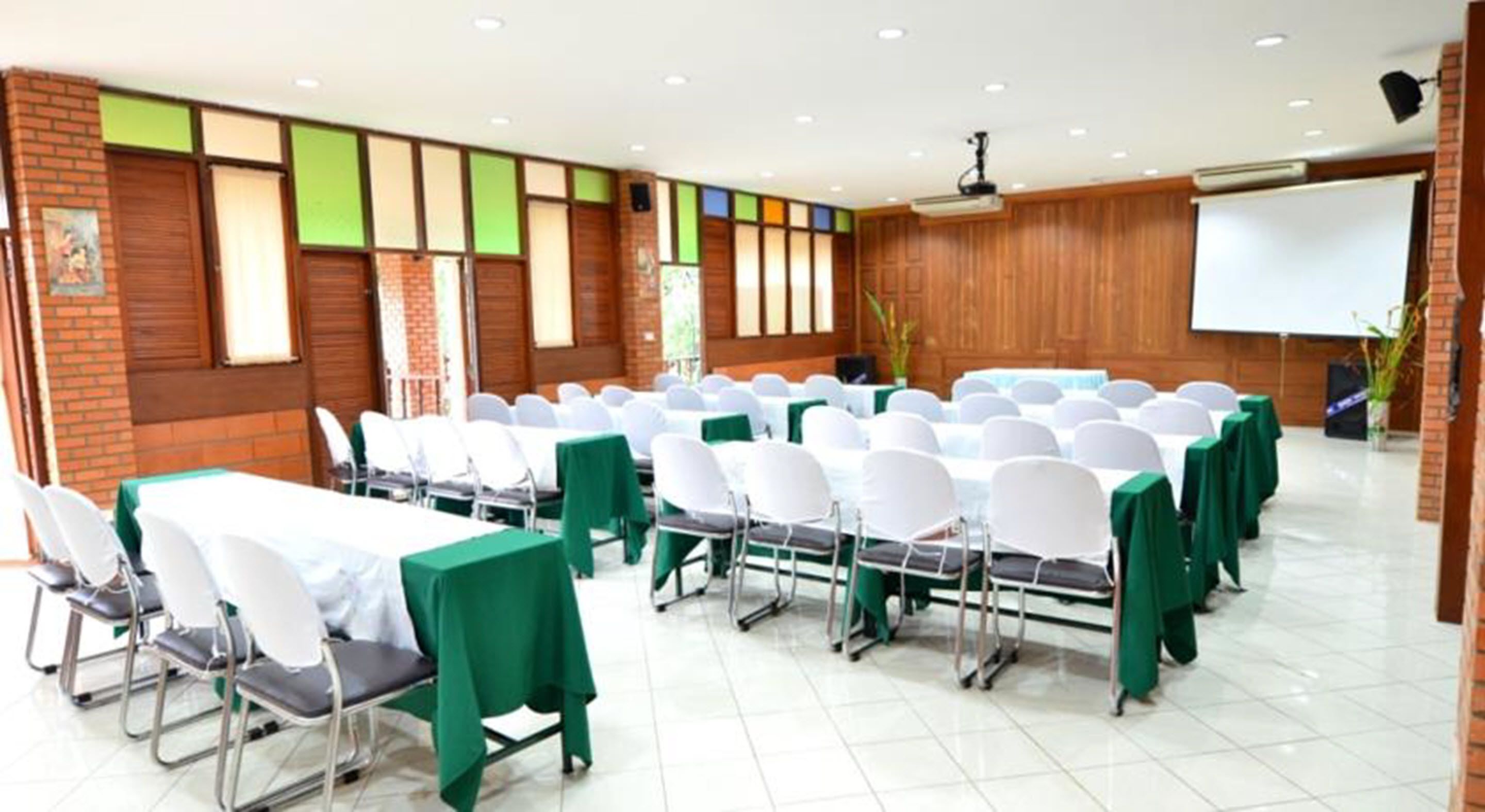 Un salón de conferencias con filas de mesas y sillas, una pantalla de proyector y una pared de ladrillo con un diseño colorido en la ventana.