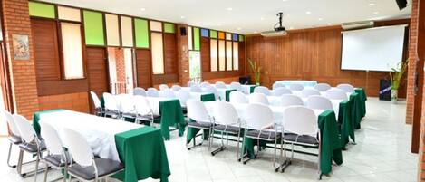 Un salón de conferencias con filas de mesas y sillas, una pantalla de proyector y una pared de ladrillo con un diseño colorido en la ventana.