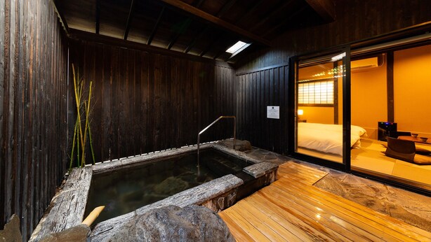 A traditional Japanese onsen with a wooden interior, a hot spring bath, and a wooden deck leading to a room with a bed and a television. - Ryokan Nagomitsuki (Beppu)