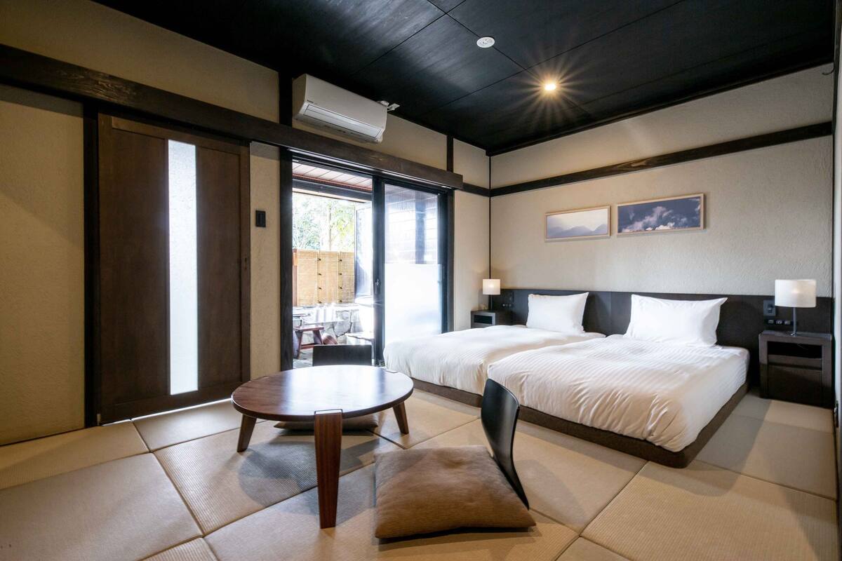 A hotel room with two beds, a dining table, and a view of the outside.