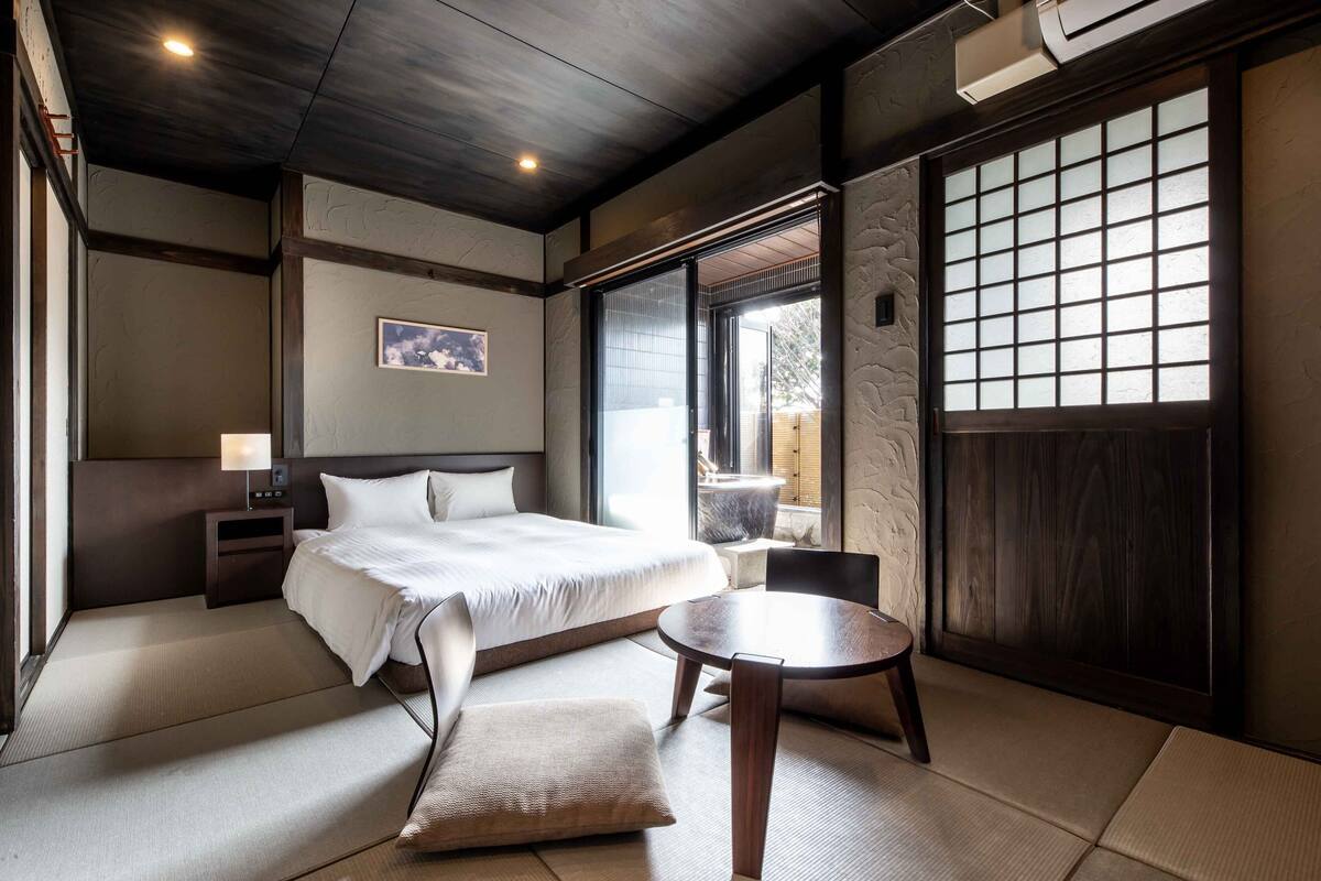 A modern hotel room with a bed, bedside table, lamp, and a view of the outdoors through a sliding door.