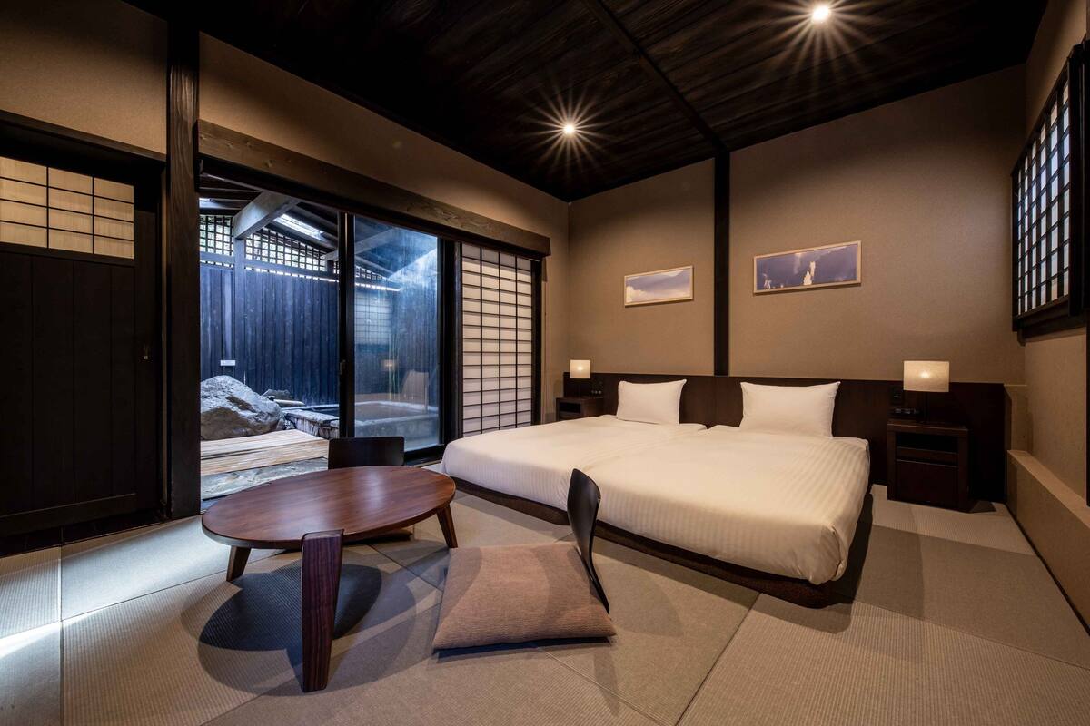 A modern hotel room with a large bed, a round dining table, and sliding doors leading to a balcony.