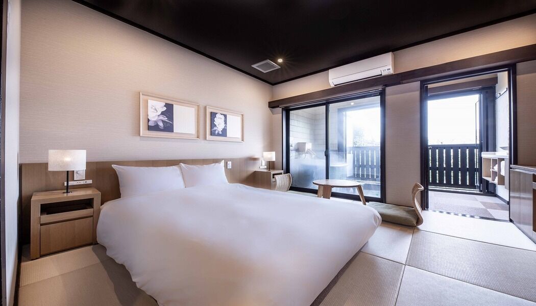 Quarto de hotel com uma cama grande, mesas de cabeceira, uma mesinha e uma varanda com vista.