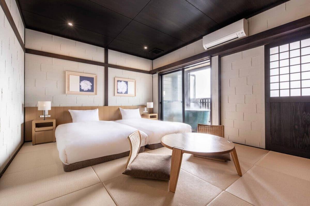 A hotel room with two beds, a small table, and a view of the outside.