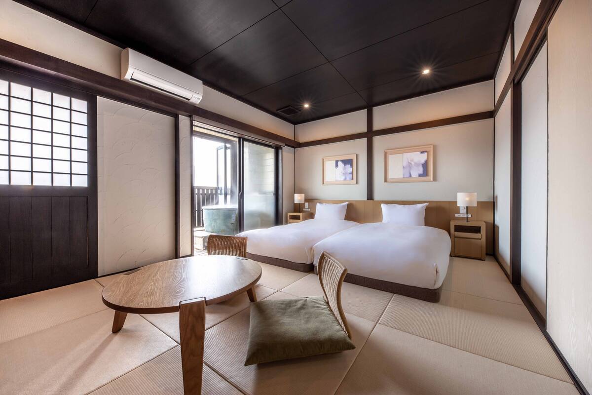 A hotel room with tatami flooring, two beds, a dining table, and a view of the outside.