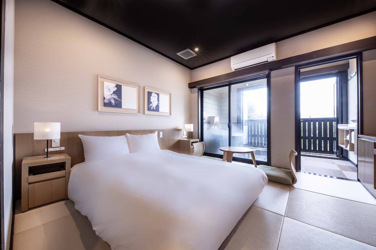 A modern hotel room with a large bed, a bedside table with a lamp, a small round table, and a view of the outside balcony.