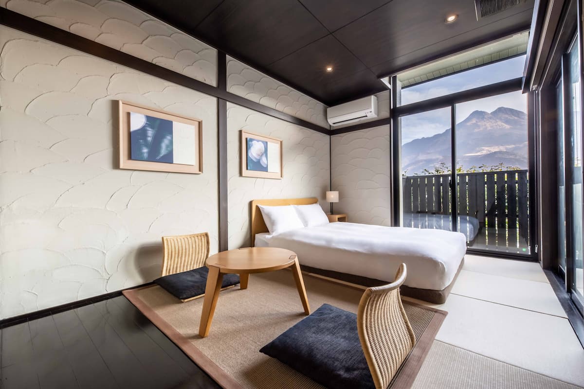 A modern hotel room with a large bed, two chairs, a small table, and a view of mountains through large windows.