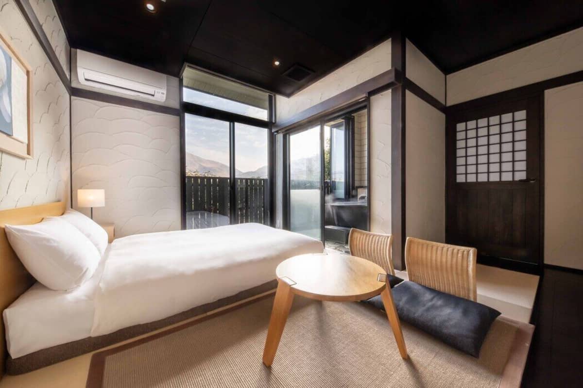 A hotel room with a bed, a round table, a chair, a view through the window, and a traditional Japanese sliding door.