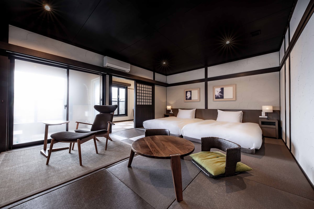 A hotel room with two beds, a sitting area with a table and chairs, and a large window providing natural light.