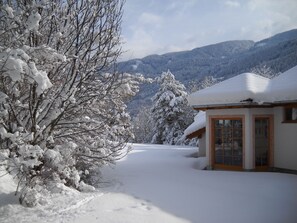 A snow-covered landscape with a building featuring a wooden door and windows, surrounded by snow-laden trees and mountains in the background. - Castelir Suite Hotel (Panchia)