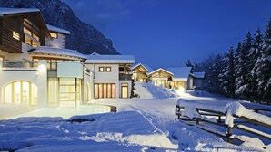 Un complejo turístico en la montaña cubierto de nieve, con varias construcciones, incluyendo una casa grande con balcón y una estructura más pequeña con techo inclinado.