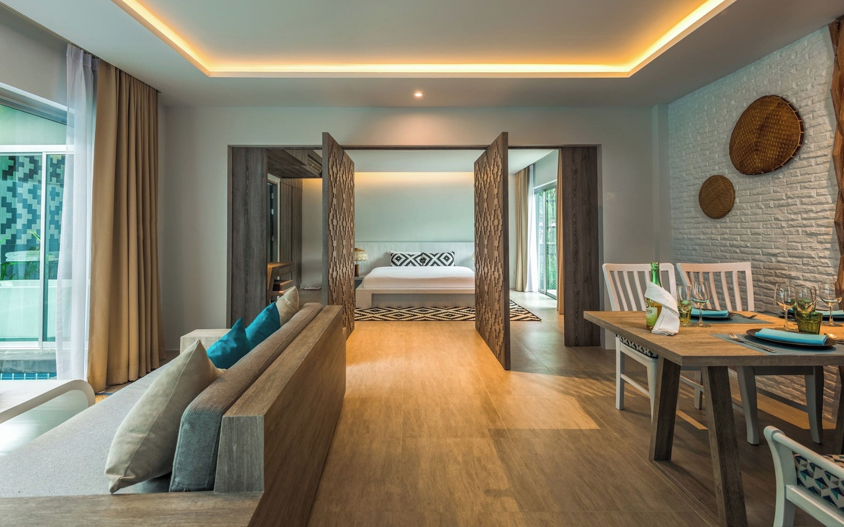 A modern hotel room with a wooden floor, a sofa with cushions, a dining table set for two, and a bed with a patterned pillow.