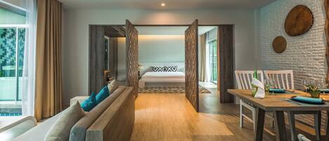 A modern hotel room with a wooden floor, a sofa with cushions, a dining table set for two, and a bed with a patterned pillow.