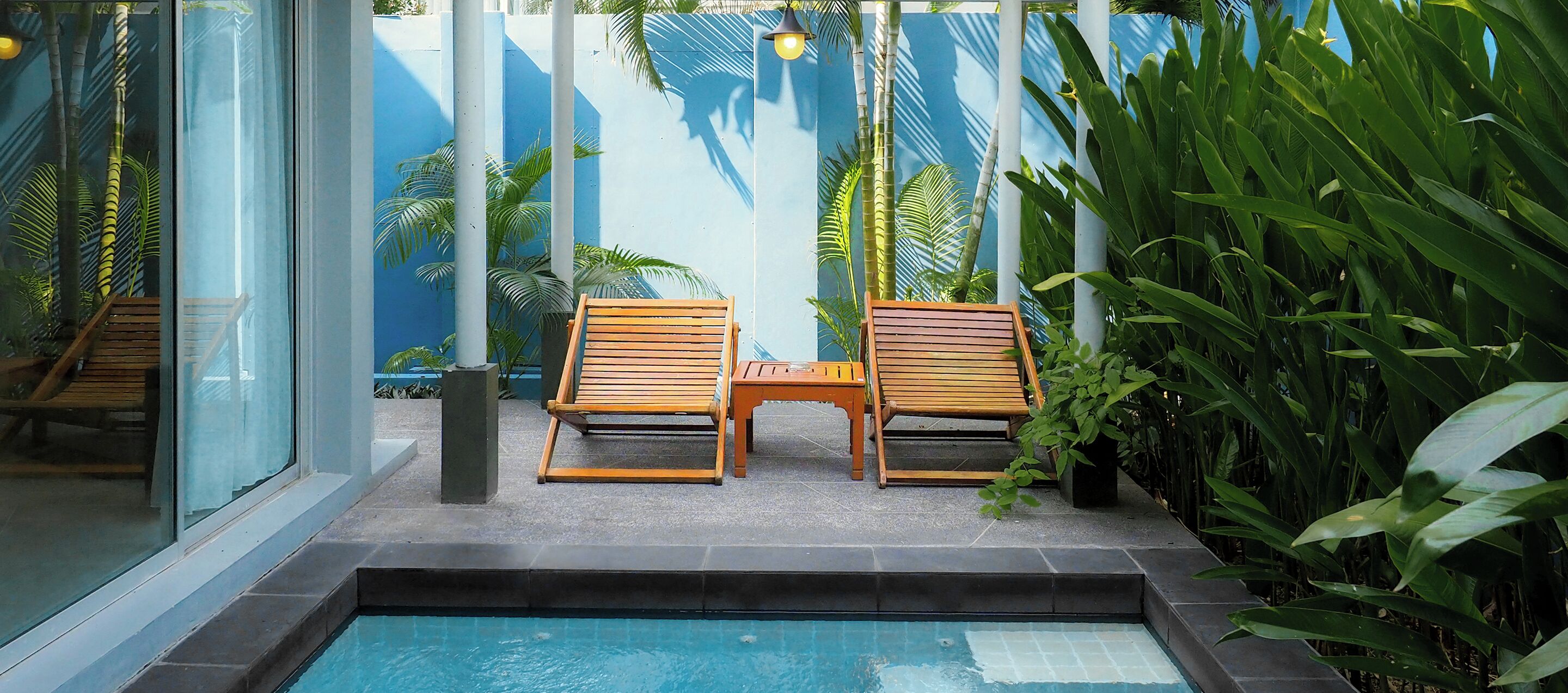 Un área junto a la piscina con dos sillones de madera y una mesita, rodeada de exuberante follaje tropical y una pared azul.