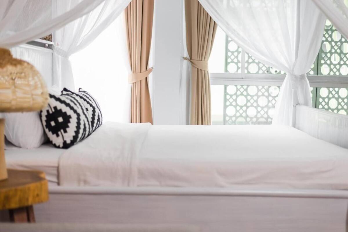 Uma cama com roupa de cama branca, uma almofada estampada e cortinas leves.