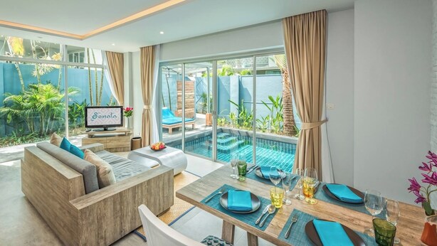 A modern living room with a sofa, dining table, and a TV. There is a pool visible through the glass doors. - Villa Sonata Phuket (Chalong)