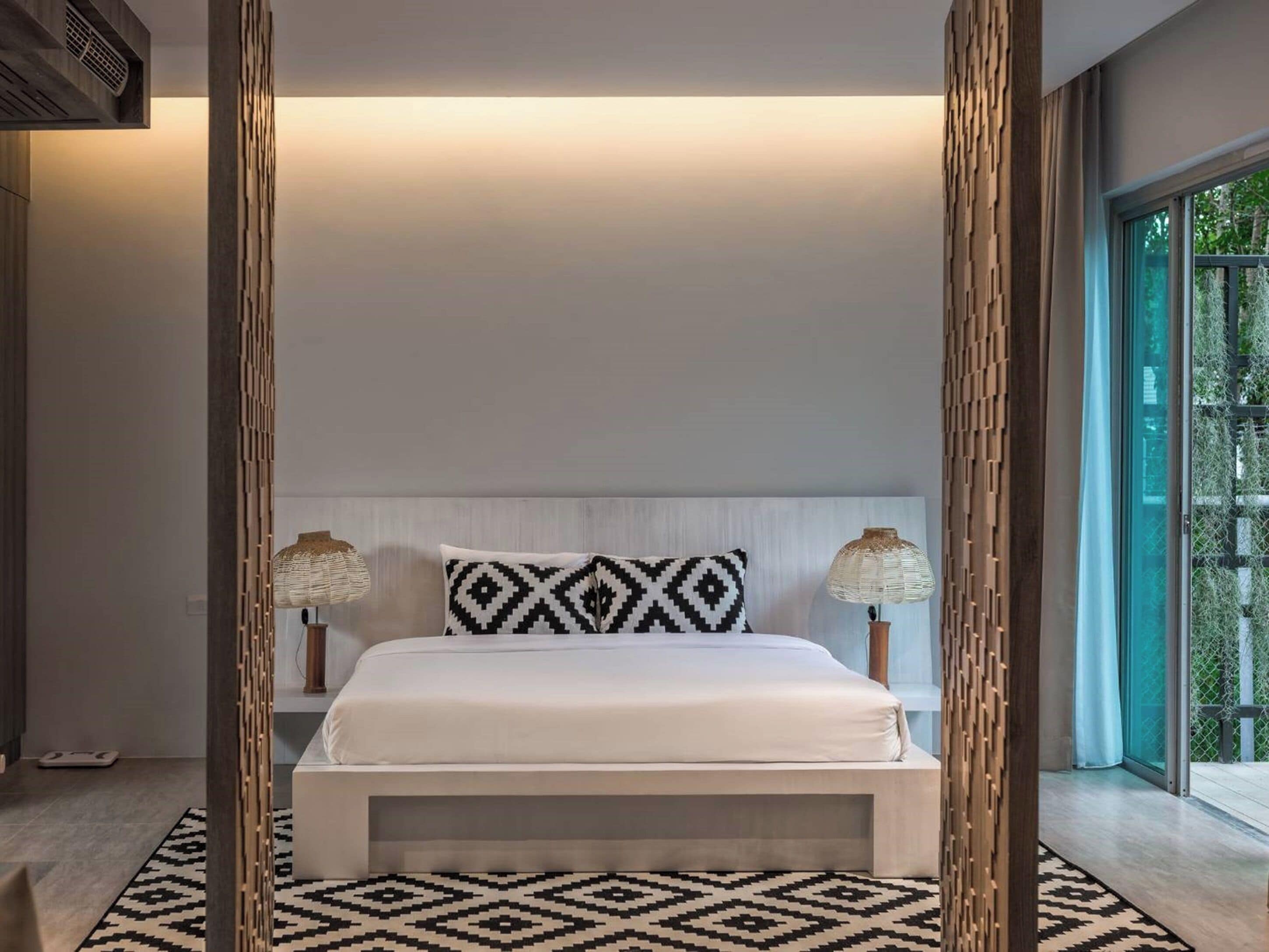 A modern bedroom with a large bed, two bedside lamps, and a view of greenery through the window.