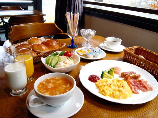 A breakfast setting with a variety of dishes including eggs, sausages, and a bowl of soup. There are also glasses of milk and orange juice, and a basket of bread.