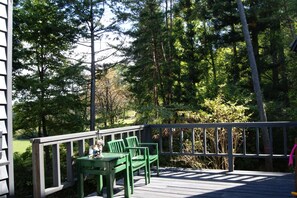 A wooden deck with green chairs and a table, surrounded by dense trees and a wooden railing.