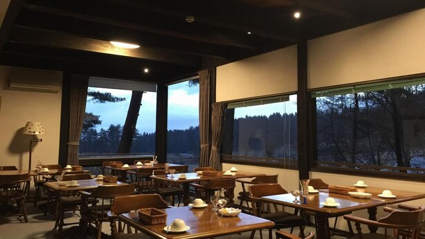 A dining area with tables set for a meal, large windows with a view of trees, and a ceiling with wooden beams. - Pension Good Luck Takayama (Takayama)