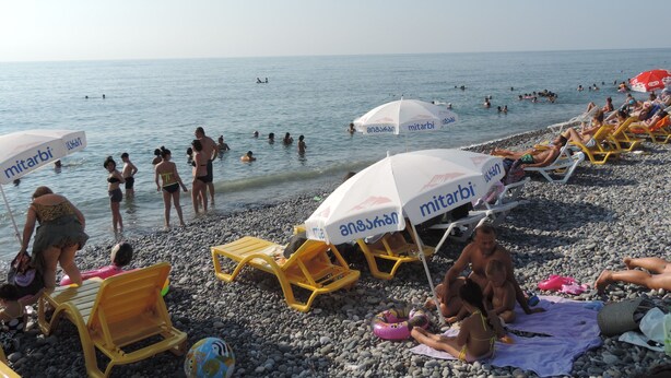 A beach with people swimming and sunbathing, yellow beach chairs, and umbrellas with 'mitarbi' branding. - Hotel Villa del Mar (Batumi)