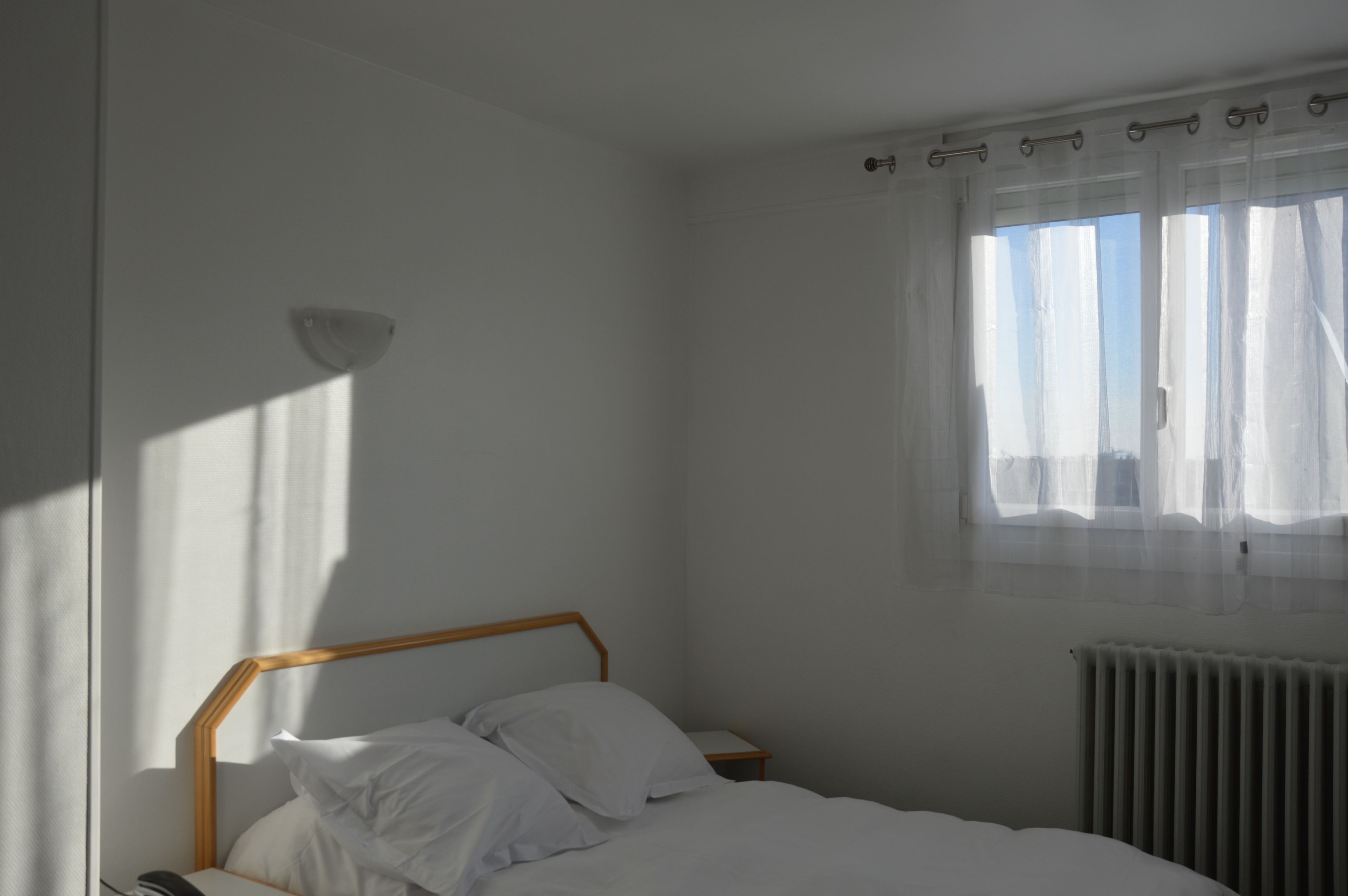 Un dormitorio con una cama individual, una mesita de noche, una lámpara de pared, un ventanal con cortinas translúcidas y un radiador.