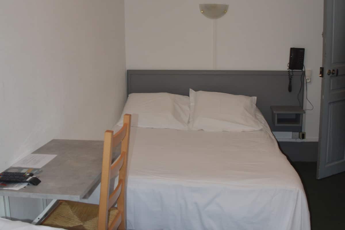 Una habitación pequeña y limpia con dos camas individuales, un escritorio, una silla, una mesita de noche, una lámpara y un teléfono.