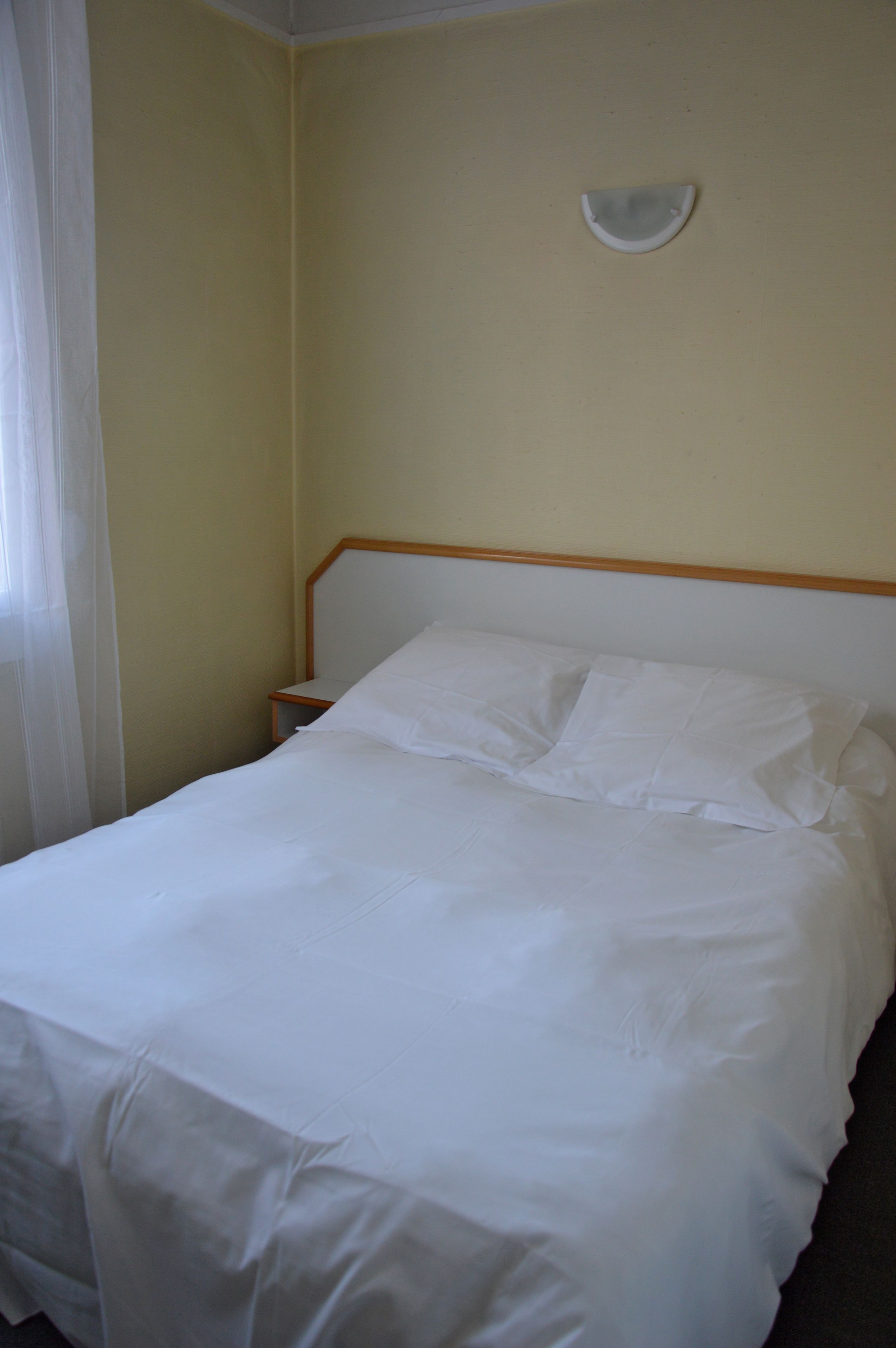 A neatly made bed with white linens in a room with a plain yellow wall and a wooden headboard.