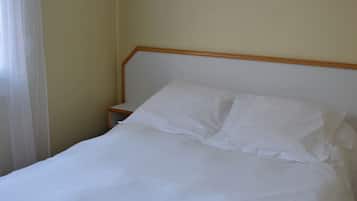 A neatly made bed with white linens in a room with a plain yellow wall and a wooden headboard.
