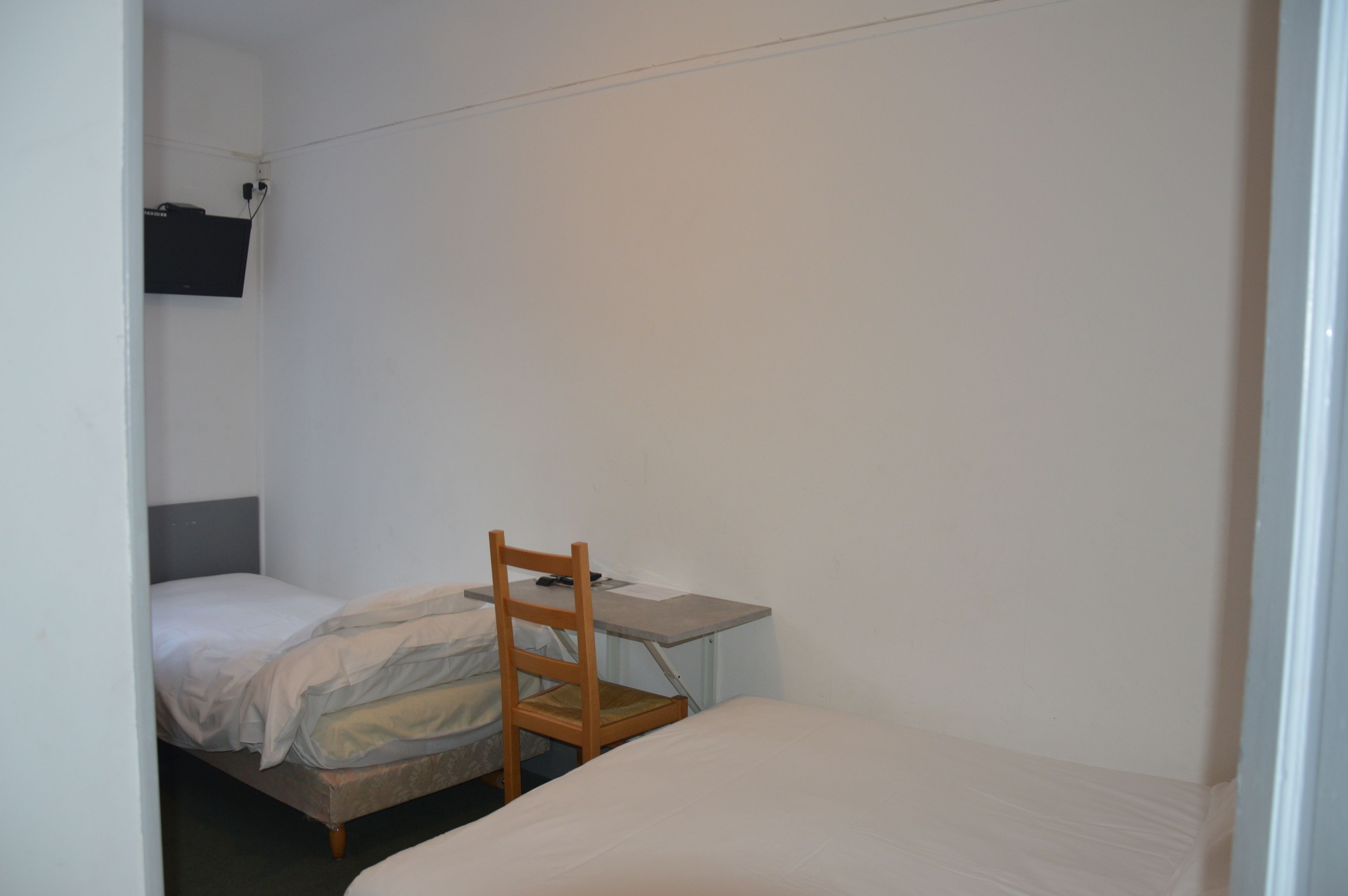 Een kleine, eenvoudig ingerichte kamer met een enkel bed, een houten stoel en een bureau.