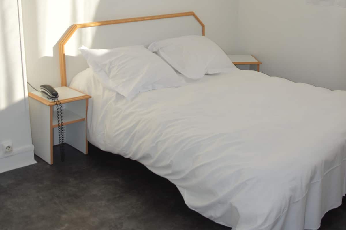 Habitación individual con una cama de una sola plaza, cabecera de madera, ropa de cama blanca, mesita de noche con lámpara y teléfono.