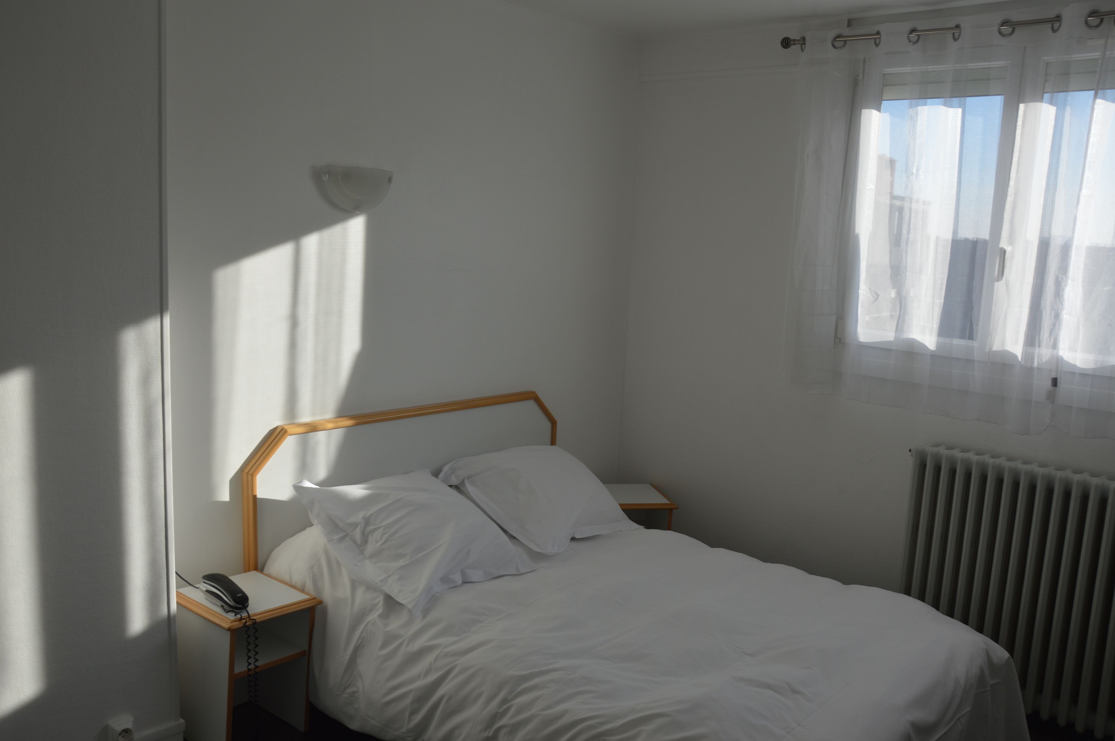 A single bed with a wooden headboard, a bedside table with a telephone, and a window with sheer curtains.