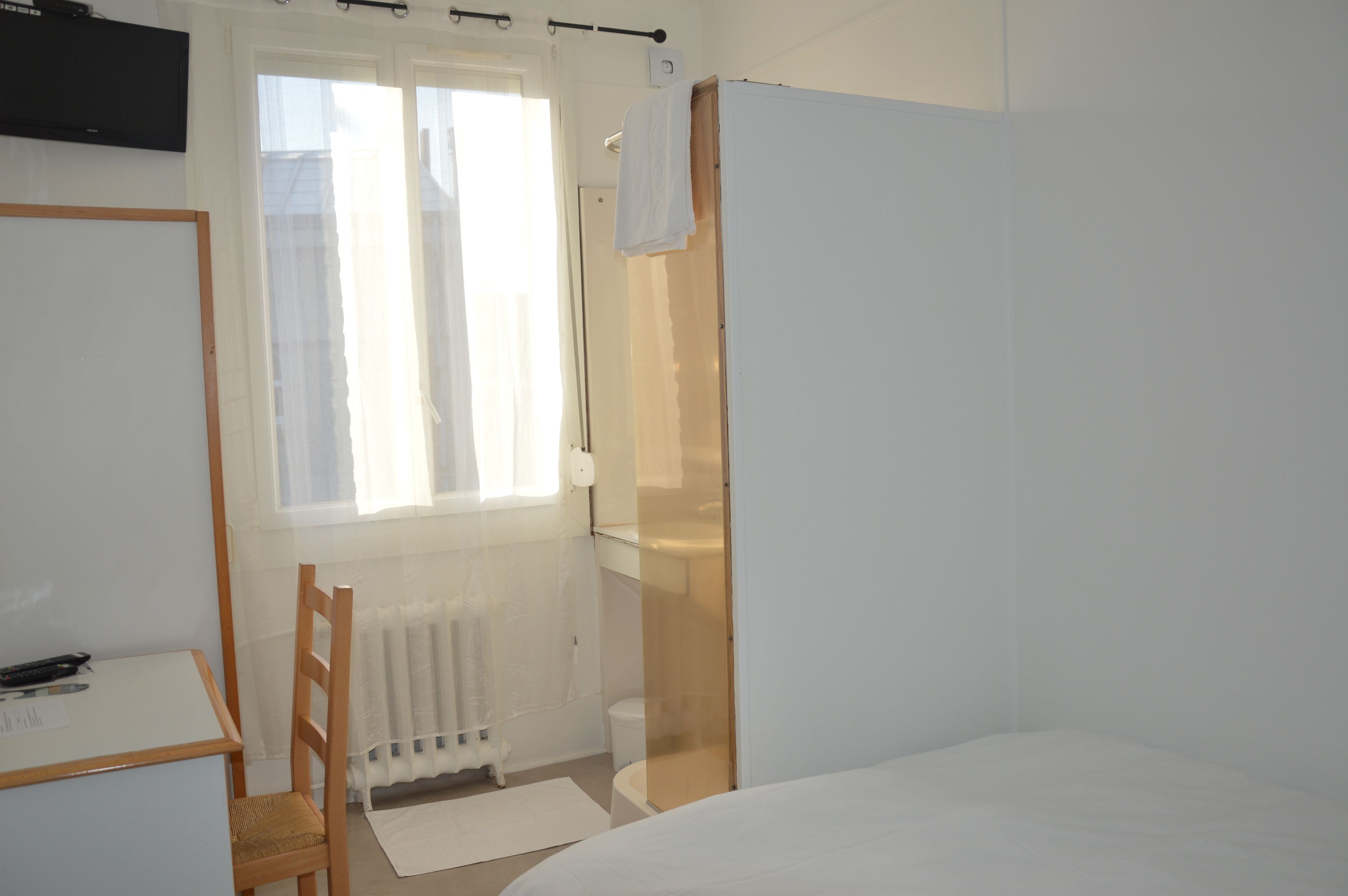 A bedroom with a bed, a chair, a radiator, a window with curtains, and a television mounted on the wall.