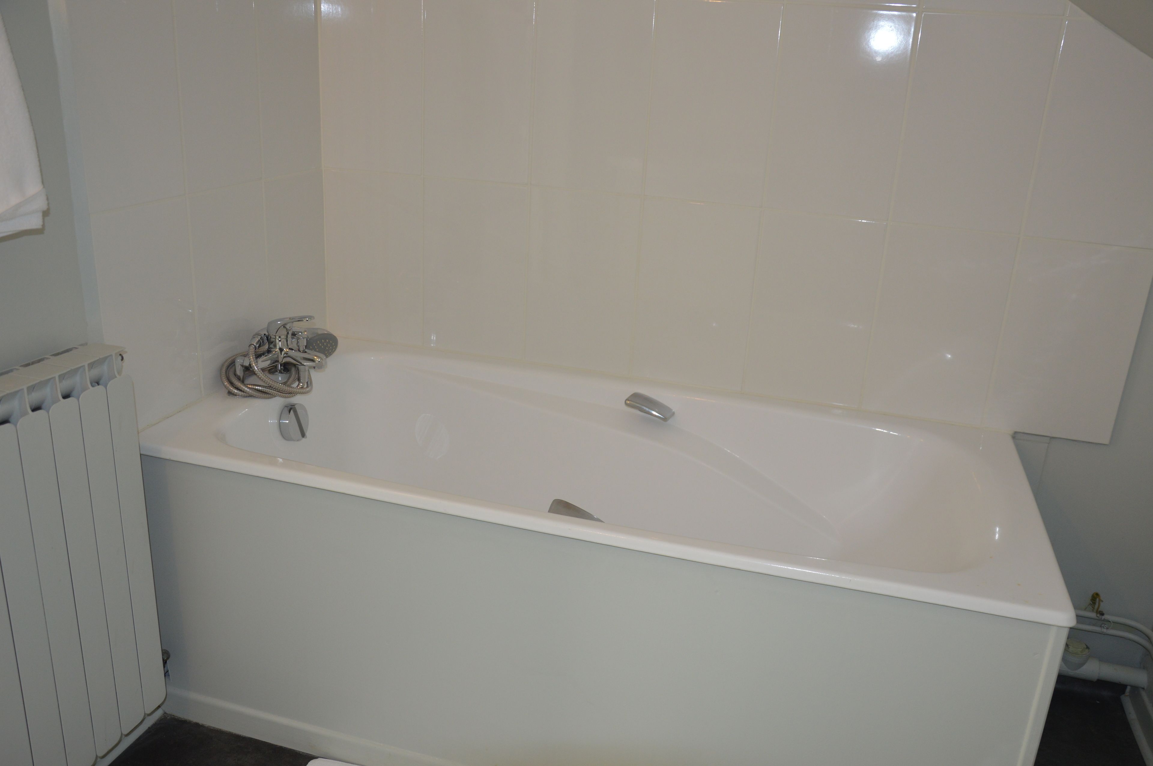 A white bathtub with a showerhead and faucet, a white radiator, and a white tiled wall.